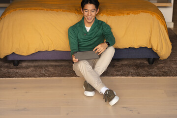 Asian man sitting on dark rug by bed with mustard yellow duvet in bedroom holding tablet
