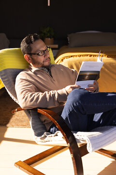 Asian man reclining on lounge chair in bedroom reading booklet beside mustard bed and potted plant
