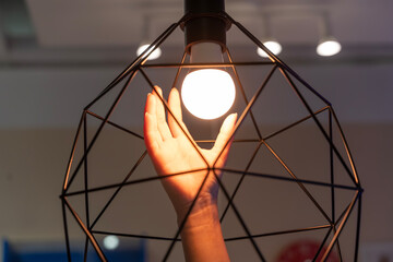 Hand touching warm light bulb inside geometric wire lamp shade with soft glow in modern interior room
