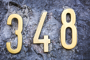 Close-up of brass house number 348 mounted on a rugged stone wall. The golden digits contrast...