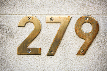 House number 279 in vintage brass mounted with visible screws on a lightly textured stone wall. The...