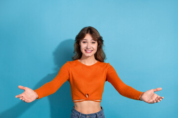 Young stylish woman in orange top smiling with open arms on a blue background, depicting happiness...