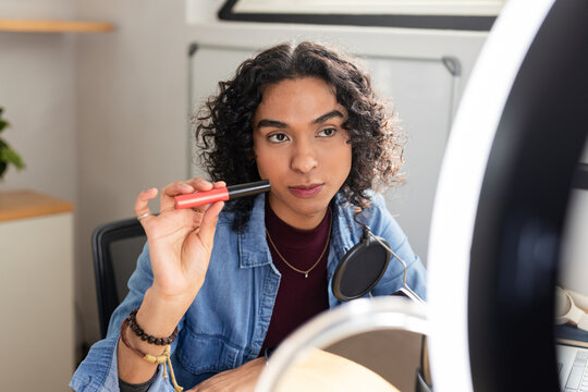 Non-binary adult holding red and black makeup tube at home studio under ring light with mic