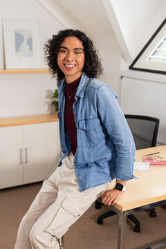 Hispanic non-binary professional leaning on wooden desk in loft-style home office with potted plant