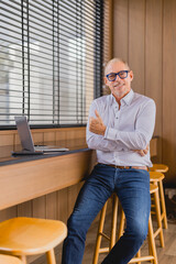 Successful senior manager showing thumbs up gesture while sitting on a stool in a modern office...