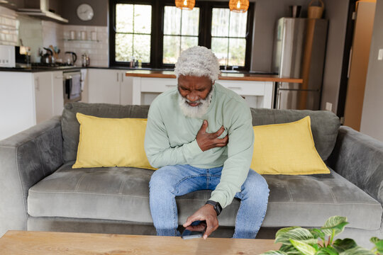 Senior African American man clutching chest reaching smartphone on coffee table in modern kitchen - Powered by Adobe
