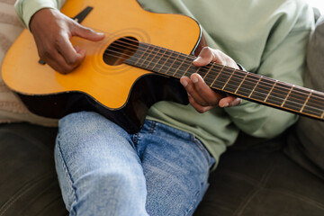 Senior African American man playing acoustic guitar while sitting on living room sofa with cushions