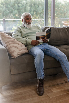 Senior African American man sitting on gray sofa in living room reading sunset brochure