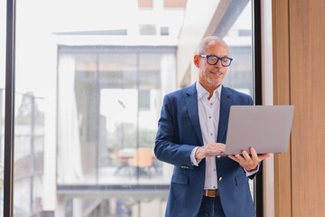 Smiling senior businessman in an elegant suit and glasses, standing by the window in a modern office, working intently on a laptop while analyzing information on the screen