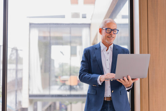Smiling senior businessman in an elegant suit and glasses, standing by the window in a modern office, working intently on a laptop while analyzing information on the screen - Powered by Adobe