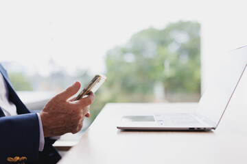 Close up of senior executive using smartphone with laptop on desk in modern office with green view...