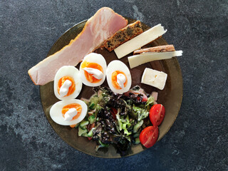 Keto breakfast, boiled eggs, bacon, cheese slices and fresh vegetables on a plate, top view