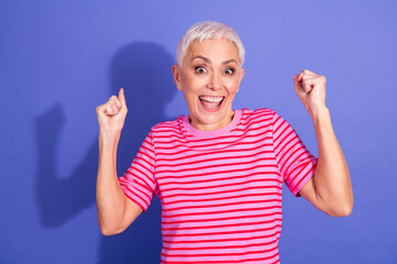 Excited senior woman celebrating success while showing cheerful gestures against a vibrant purple...