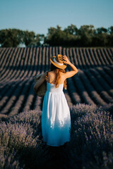 Woman walking through lavender field at sunset wearing white dress and straw hat