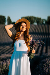 Young woman enjoying lavender field at sunset wearing white dress and straw hat