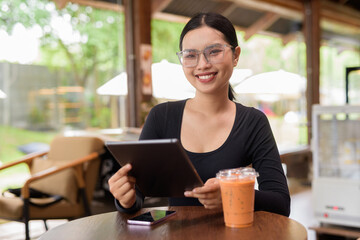 Young beautiful Asian woman working with tablet computer and drinking iced Thai tea in cafe