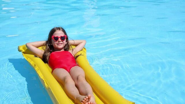 Little girl in red swimsuit lies comfortably on inflatable mattress in swimming pool with blue water on warm summer day on tropical vacation. Relaxation, fun, carefree childhood concept. Footage - Powered by Adobe
