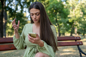 Stressed beautiful woman looking at smartphone with unpleasant surprise outdoors in park, unhappy...