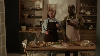 Mature diverse couple dressed in aprons drinking red wine while making pizza at wooden table in kitchen - Powered by Adobe