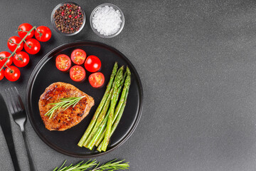 Tasty grilled beef steak served with green asparagus and cherry tomatoes on black plate on gray textured backgound. Top view, copy space. Perfect for food, diet, or culinary design concepts.