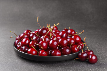 Close up of black plate filled with ripe red sweet cherry placed on dark gray textured background. Delicious food, juicy summer fruit and healthy eating.