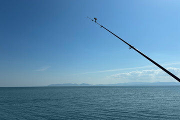 Peaceful sea fishing with a rod against clear blue sky and calm ocean water, tranquil coastal landscape with distant mountains, summer outdoor leisure and recreational activity concept.