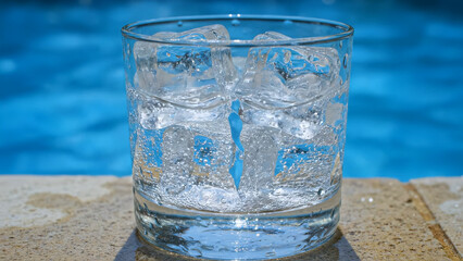 Glass filled with ice cubes sits in front of shimmering water in swimming pool, offering visual sense of refreshment and cool relief. Crisp sparkle of ice, perfect antidote to summer heat and thirst