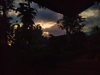 A dramatic sunset or sunrise sky with dark, heavy clouds and a sliver of golden light on the horizon, silhouetting tropical trees.
