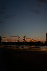 A serene twilight scene features a lit bridge over still waters, a crescent moon in the tranquil...