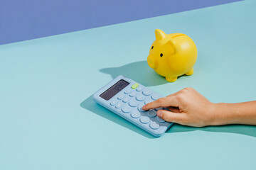 Hand using calculator next to yellow piggy bank on blue background with copy space