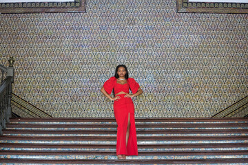 Young, beautiful, African woman, wearing a red dress, posing as a model with her hands on her waist on the stairs of the square of Spain in Seville. Fashion concept, beauty, trendy.
