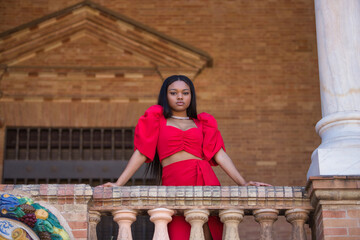 Young, beautiful, African woman in a red dress, with a serious gesture looking at the camera leaning on a railing in the square of Spain in Seville. Fashion concept, beauty, trendy.