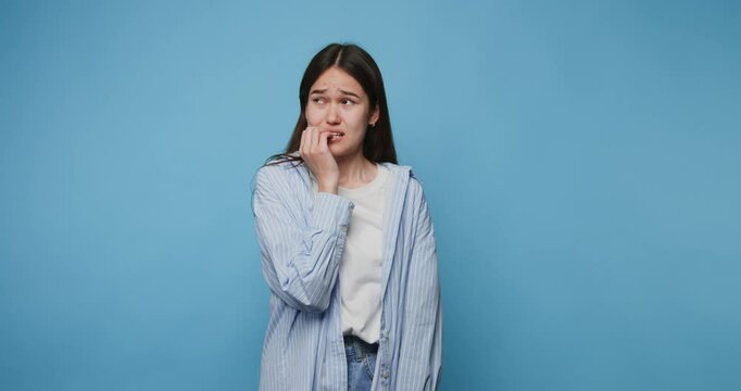 Young woman expresses concern against a blue background while biting her fingernails