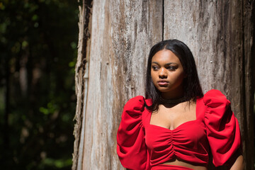 Young, beautiful, African woman in a red dress, posing leaning on the trunk of a large tree in connection with nature. Concept fashion, beauty, trendy.