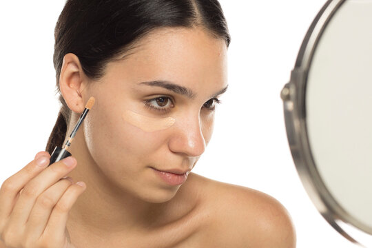 Young woman applying concealer under eye while looking in mirror, studio shot on white background