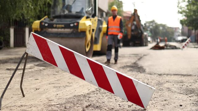 Workers are busy on a road construction site with heavy machinery operating nearby. Safety barriers mark the area, ensuring pedestrian guidance amidst the ongoing work.
