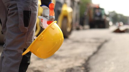 A construction worker stands on a busy worksite holding a yellow hard hat while preparing for road repairs. Heavy machinery is visible in the background as the day progresses.
