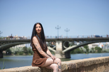 Young woman, brunette, tall, thin and very tattooed, with brown dress, looking to infinity sitting by the river with the Triana bridge in the background. Concept of youth, millennial.