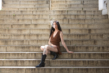 Young woman, brunette, tall, thin and very tattooed, with brown dress and boots, sweet, innocent and lonely, vaping, sitting on some stairs. Concept of youth, beauty, rebelliousness, millennial.