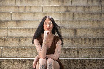 Young woman, dark-haired, tall, thin, and very tattooed, wearing a brown dress, with her hand on her chin, looking at the camera, sweet, innocent, and lonely, sitting on some stairs.