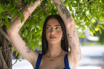 Portrait of a young woman, brunette, tall, slim, and very tattooed, wearing a blue top, with a distracted gaze holding onto a vine of green leaves. Concept of youth, beauty, rebellion, millennial.