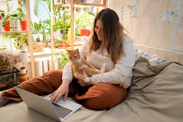 Plus size woman sits on her bed with a laptop, working remotely while holding her cute orange cat....