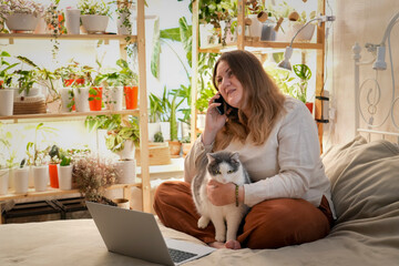 A plus size model enjoys a relaxing day working remotely in her vibrant bedroom, talking on the phone while her cat sits comfortably in her lap, surrounded by lush greenery