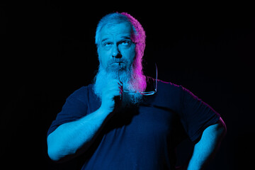 Pensive bearded man holding his glasses, deep in thought, with a moody blue and pink neon light backdrop.