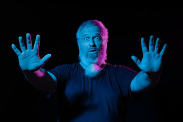 A man with a surprised expression, raising his hands, lit by blue and pink neon lights against a dark background.
