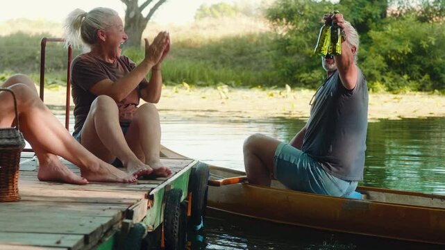 An excited 50s man in a canoe surprises his friends by pulling a chilled beer from the river during a summer river party on a dock by the river.