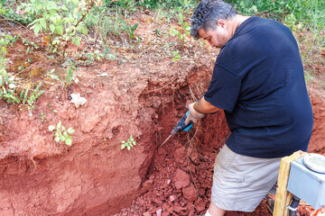 Individual works diligently in rich red soil, using jackhammer tool to break up ground