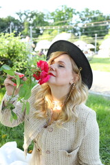 A woman in a stylish outfit sits in a vibrant park, inhaling the aroma of fresh roses. Sunlight...