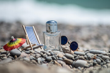 Perfume bottle next to sun lounger, umbrella and sunglasses on pebbles with sea in background....