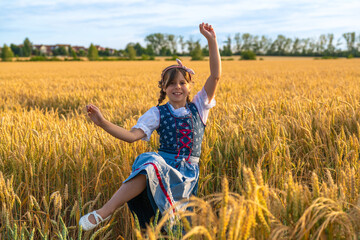 Joyful child in traditional Bavarian dirndl dress playing in a wheat field. Raised arms and leg...